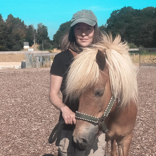 Laure de l'association Libea pose avec un cheval islandais alezan clair à crinière blonde, en licol, dans une carrière extérieure entourée d'arbres.
