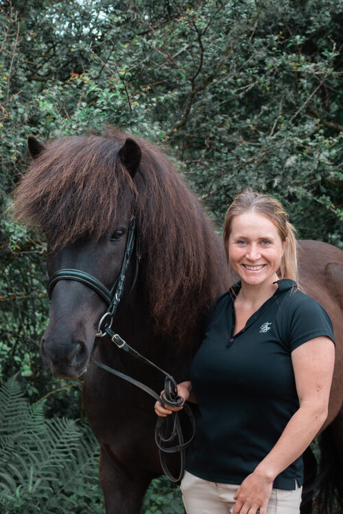 Lisa de l'association Libea pose avec un cheval islandais noir aux crins fournis, tenu en longe, devant une haie d'arbustes.