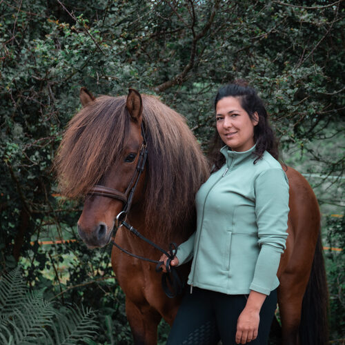 Tabea de l'association Libea pose avec un cheval islandais bai aux crins abondants, en filet, devant un décor boisé et verdoyant.