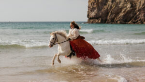 Lisa de l’association Libea monte Glanni Glæpur frá Breiðumörk 2, cheval islandais blanc, au galop dans les vagues sur une plage bretonne, vêtue d’une robe longue rouge et blanche.