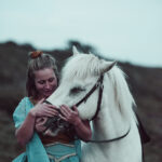 Lisa de l’association Libea enlace tendrement Glanni Glæpur frá Breiðumörk 2, cheval islandais blanc, dans une tenue colorée en extérieur au crépuscule.