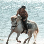 Lisa de l'association Libea monte Glanni Glæpur frá Breiðumörk 2, cheval islandais blanc, au galop sur la plage, longeant l’eau scintillante.