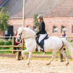 Lisa de l'association Libea monte Glanni Glæpur frá Breiðumörk 2, cheval islandais blanc, en concours dans une carrière extérieure devant un bâtiment en briques.