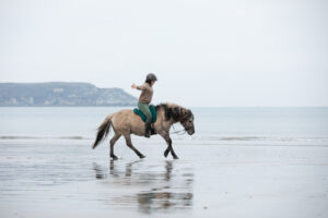 Tabea de l'association Libea galopant librement les bras ouverts sur la plage avec Phönix, cheval islandais gris souris