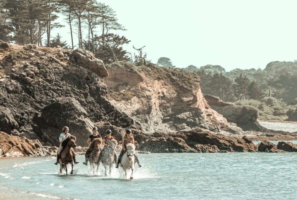Quatre cavalières de l’association Libea galopent dans l’eau sur leurs chevaux islandais, longeant les rochers escarpés d’une crique bretonne sous un ciel clair.