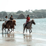 Trois cavalières de l’association Libea avancent au pas sur leurs chevaux islandais en longeant le rivage, la mer scintillant sous le soleil et les falaises bretonnes en arrière-plan.
