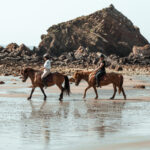 Deux cavalières de l’association Libea chevauchent leurs chevaux islandais sur le sable humide, au pied des falaises rocheuses bretonnes, lors de la prestation Mer & Bruyère.